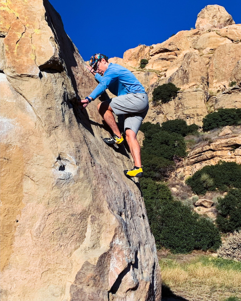 A man in a blue shirt and yellow climbing shoes climbs up the side of a large boulder at Stony Point